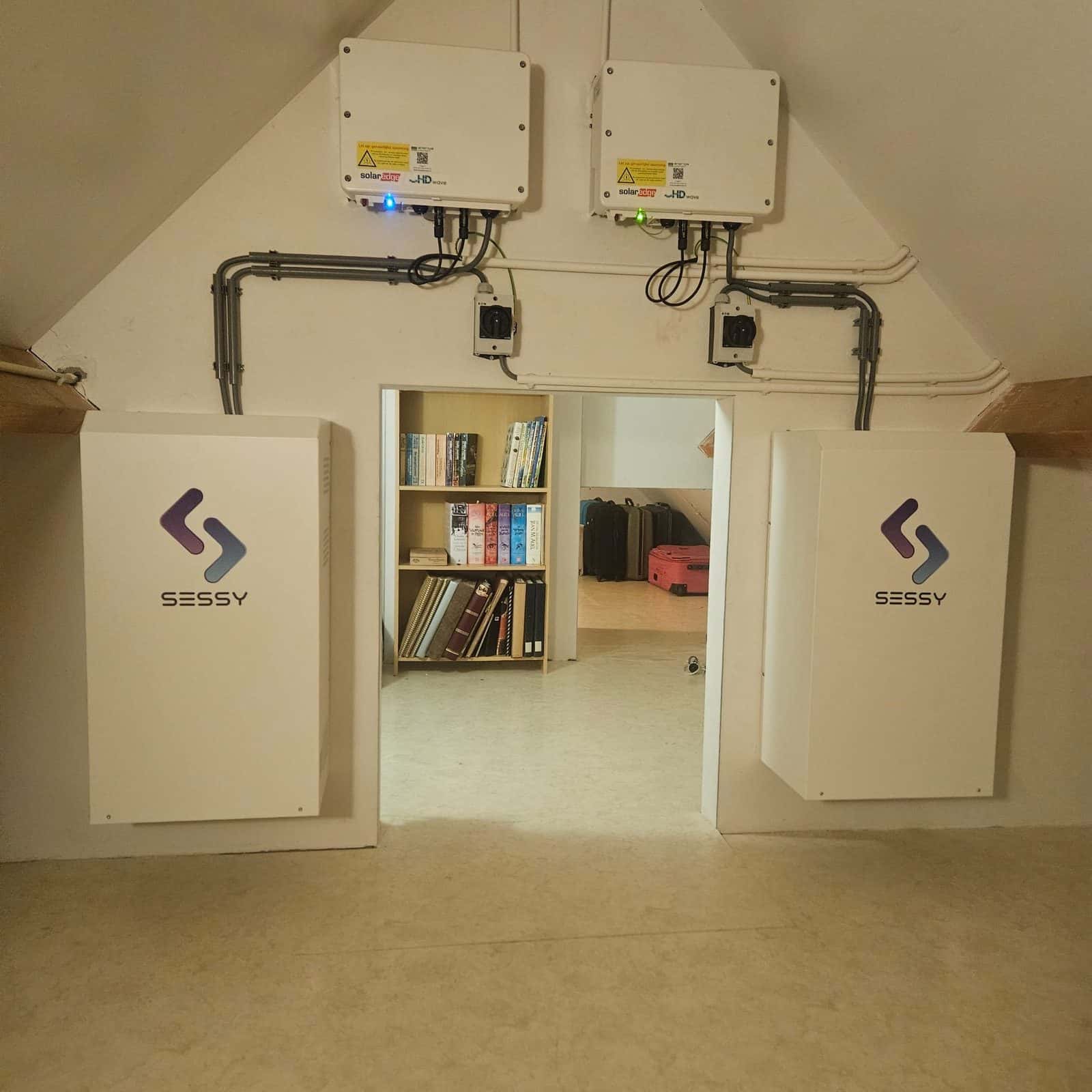 Twee Sessy thuisbatterijen aan de wand op een zolder, met de omvormers erboven
